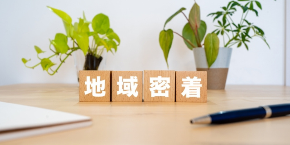 『地域密着』の文字が書かれた木のブロックと観葉植物、ペンのあるデスク