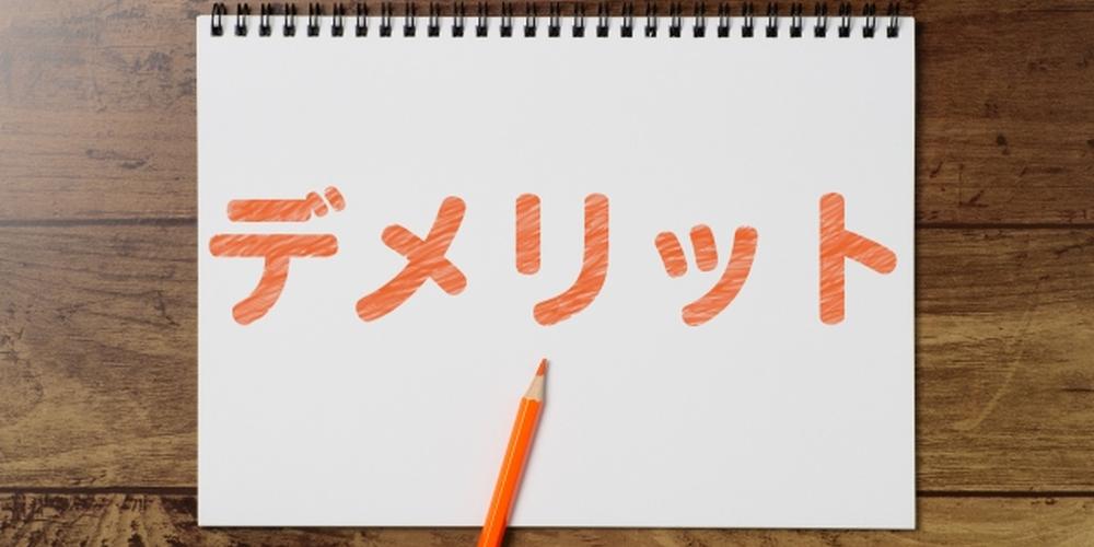 「デメリット」とオレンジ色で書かれたスケッチブックとオレンジの色鉛筆が並ぶ木目調の机上イメージ