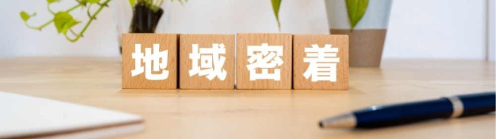 “地域密着”の文字が書かれた木製ブロックと観葉植物が並ぶデスクの上の風景。地元に根ざしたサービスの象徴
