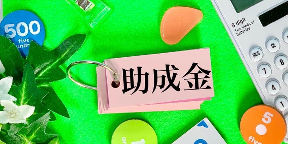 「助成金」と書かれたタグと電卓、コインが置かれた緑色の背景