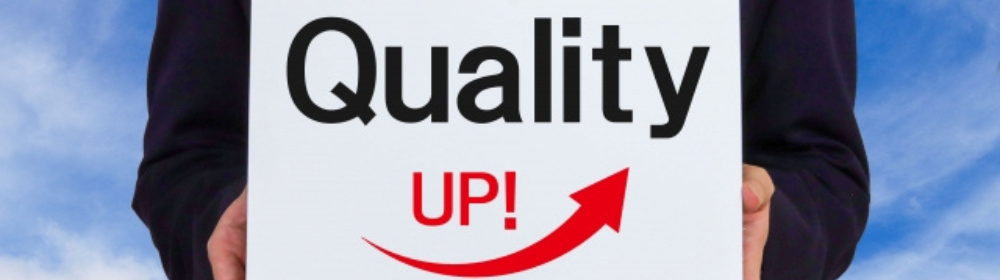 「Quality UP！」と書かれた看板を掲げるスーツ姿の人物