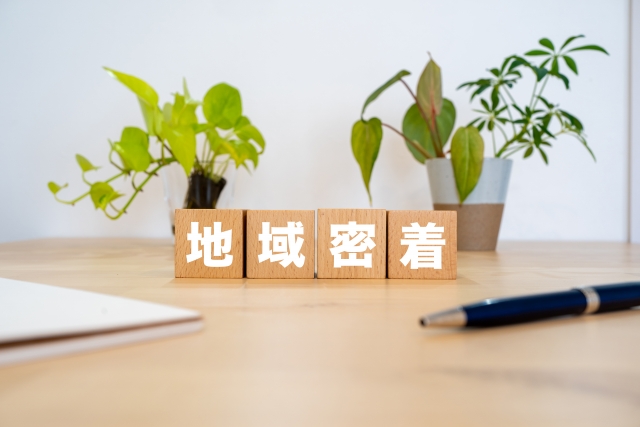 “地域密着”と書かれた木製ブロックが観葉植物と一緒にテーブルに置かれたイメージ。地域との信頼関係や密接なつながりを表すビジュアル。