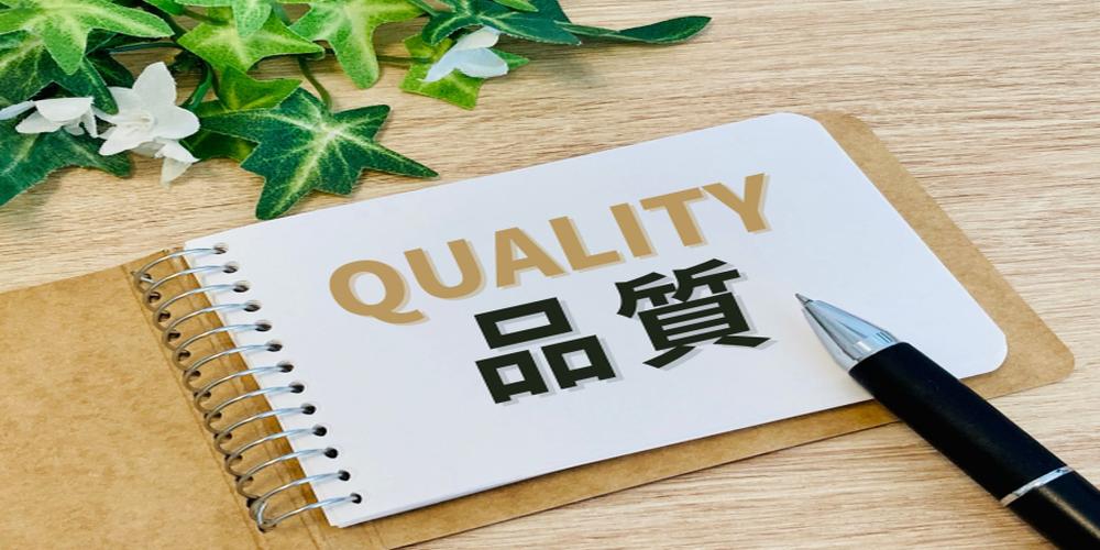 “QUALITY”と“品質”の文字がノートに書かれたイメージ。ペンと観葉植物が添えられ、製品やサービスの高品質を印象づけるビジュアル。